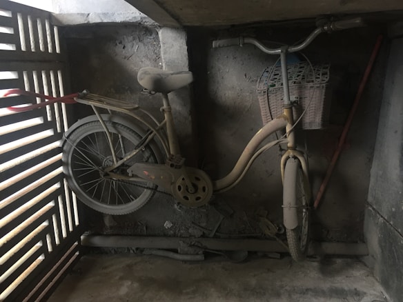 A dusty, old bicycle is leaning against a rough concrete wall in a dimly lit space. The bicycle has a rusty brown hue, and its basket is partially woven. A wooden slatted panel to the left allows some light to filter in, casting shadows on the bike. Debris and some pipes are visible on the floor.