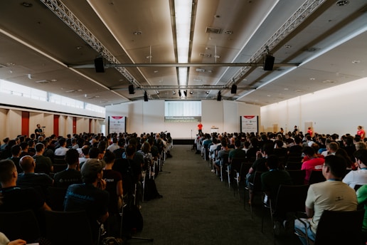 A large room filled with people seated and facing a stage where a speaker is presenting. The room has a modern design with a high ceiling and is well-lit with natural light from the windows on the left. The audience appears attentive, and some individuals are standing on the sides.