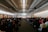 A large room filled with people seated and facing a stage where a speaker is presenting. The room has a modern design with a high ceiling and is well-lit with natural light from the windows on the left. The audience appears attentive, and some individuals are standing on the sides.