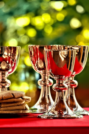 Several silver chalices with elegant stems are neatly arranged against a softly blurred background of green and yellow hues. A stack of flat, golden-brown wafers is placed on a tray nearby, resting on a rich red surface.