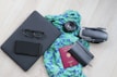 A neatly arranged collection of travel essentials on a light wooden floor, featuring a black laptop with eyeglasses and a smartphone on top, a vibrant blue and green patterned scarf, a camera lens, a folded drone, a closed black notebook, and a red passport.