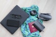 A neatly arranged collection of travel essentials on a light wooden floor, featuring a black laptop with eyeglasses and a smartphone on top, a vibrant blue and green patterned scarf, a camera lens, a folded drone, a closed black notebook, and a red passport.