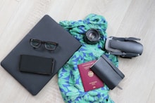 A neatly arranged collection of travel essentials on a light wooden floor, featuring a black laptop with eyeglasses and a smartphone on top, a vibrant blue and green patterned scarf, a camera lens, a folded drone, a closed black notebook, and a red passport.