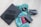 A neatly arranged collection of travel essentials on a light wooden floor, featuring a black laptop with eyeglasses and a smartphone on top, a vibrant blue and green patterned scarf, a camera lens, a folded drone, a closed black notebook, and a red passport.
