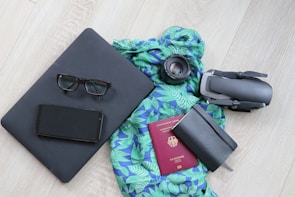 A neatly arranged collection of travel essentials on a light wooden floor, featuring a black laptop with eyeglasses and a smartphone on top, a vibrant blue and green patterned scarf, a camera lens, a folded drone, a closed black notebook, and a red passport.