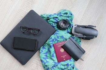 A neatly arranged collection of travel essentials on a light wooden floor, featuring a black laptop with eyeglasses and a smartphone on top, a vibrant blue and green patterned scarf, a camera lens, a folded drone, a closed black notebook, and a red passport.
