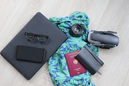 Uma coleção organizada de itens essenciais de viagem disposta em um piso de madeira clara, apresentando um laptop preto com óculos e um smartphone por cima, um cachecol vibrante com estampa azul e verde, uma lente de câmera, um drone dobrado, um caderno preto fechado e um passaporte vermelho.