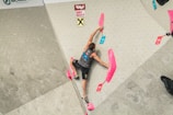 A person is climbing an indoor rock climbing wall, gripping pink climbing holds. The climber is wearing a sleeveless top and shorts, with their right arm outstretched towards the top hold. Various colored labels indicating 'top' and 'zone' are visible on the wall.