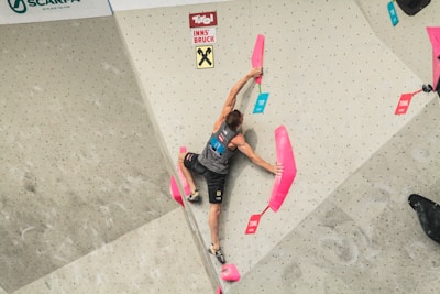 A person is climbing an indoor rock climbing wall, gripping pink climbing holds. The climber is wearing a sleeveless top and shorts, with their right arm outstretched towards the top hold. Various colored labels indicating 'top' and 'zone' are visible on the wall.