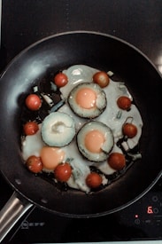 A frying pan contains sliced vegetables and eggs being cooked. The vegetables include sliced avocado rings with eggs cooked inside each, surrounded by cherry tomatoes. The scene is set on a stovetop with a digital control pad visible.