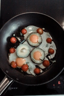 A frying pan contains sliced vegetables and eggs being cooked. The vegetables include sliced avocado rings with eggs cooked inside each, surrounded by cherry tomatoes. The scene is set on a stovetop with a digital control pad visible.