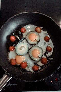 A frying pan contains sliced vegetables and eggs being cooked. The vegetables include sliced avocado rings with eggs cooked inside each, surrounded by cherry tomatoes. The scene is set on a stovetop with a digital control pad visible.