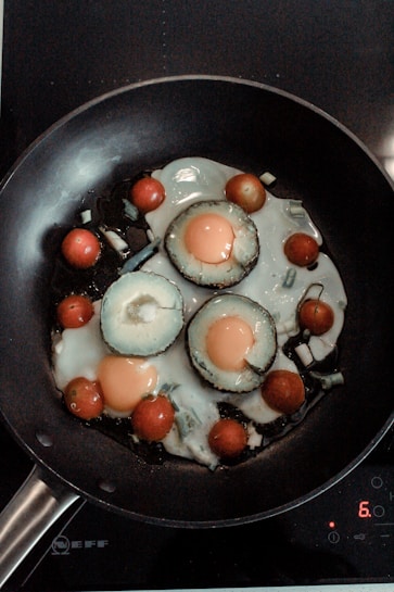 A frying pan contains sliced vegetables and eggs being cooked. The vegetables include sliced avocado rings with eggs cooked inside each, surrounded by cherry tomatoes. The scene is set on a stovetop with a digital control pad visible.