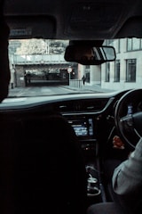 A view from inside a vehicle looking out through the windshield at a street scene. The driver is partially visible, holding the steering wheel. The dashboard and a black object, possibly a cup holder or a compartment, are in view. Outside, a road with signs indicating height restrictions leads under a railway bridge, with a pedestrian walking on the sidewalk to the right.