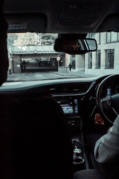 A view from inside a vehicle looking out through the windshield at a street scene. The driver is partially visible, holding the steering wheel. The dashboard and a black object, possibly a cup holder or a compartment, are in view. Outside, a road with signs indicating height restrictions leads under a railway bridge, with a pedestrian walking on the sidewalk to the right.
