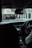 A view from inside a vehicle looking out through the windshield at a street scene. The driver is partially visible, holding the steering wheel. The dashboard and a black object, possibly a cup holder or a compartment, are in view. Outside, a road with signs indicating height restrictions leads under a railway bridge, with a pedestrian walking on the sidewalk to the right.