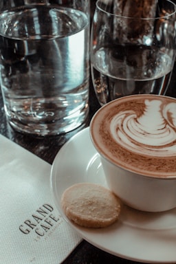 A cup of cappuccino with intricate latte art is placed on a white saucer alongside a small round cookie. Two glasses of water are in the background, and a napkin with the words 'Grand Cafe' is visible.