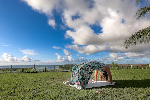 A person performs a backbend yoga pose on a lush green lawn, with a picturesque view of a distant body of water and hills under a vivid blue sky filled with scattered clouds. A palm tree is visible on the right side.