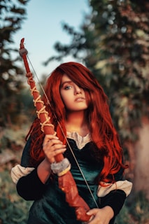 A person with long red hair holds an intricately carved wooden staff. They are dressed in a dark green outfit with light-colored accents. The background features blurred greenery, creating a natural setting.