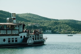 A paddle steamboat is on a calm body of water, surrounded by rolling green hills in the background. Several people are seated on the boat, enjoying the scenic view, while another boat with a sail is further away on the water. The sky is clear and bright.