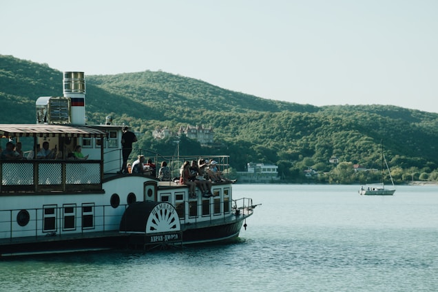 A paddle steamboat is on a calm body of water, surrounded by rolling green hills in the background. Several people are seated on the boat, enjoying the scenic view, while another boat with a sail is further away on the water. The sky is clear and bright.