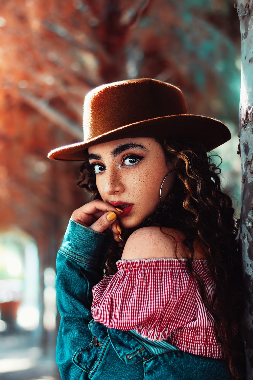 woman in red off-shoulder top and blue jacket leaning on tree trunk during daytime