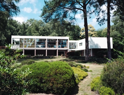 A modernist house with large windows is situated within a lush, green landscape. Tall trees frame the setting, and various shrubs and bushes are arranged around the foreground, adding to the natural atmosphere. The white facade of the building contrasts with the greenery around it.