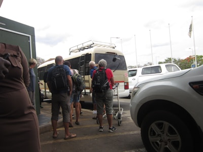 A group of people with backpacks are standing outside, waiting to board or unload a tan minibus. Several parked white vehicles are visible nearby, and some people are holding trolley luggage. The sky is overcast, and there are tall flagpoles in the background.