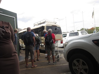 A group of people with backpacks are standing outside, waiting to board or unload a tan minibus. Several parked white vehicles are visible nearby, and some people are holding trolley luggage. The sky is overcast, and there are tall flagpoles in the background.