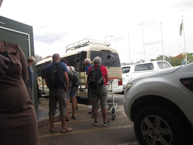 A group of people with backpacks are standing outside, waiting to board or unload a tan minibus. Several parked white vehicles are visible nearby, and some people are holding trolley luggage. The sky is overcast, and there are tall flagpoles in the background.