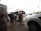 A group of people with backpacks are standing outside, waiting to board or unload a tan minibus. Several parked white vehicles are visible nearby, and some people are holding trolley luggage. The sky is overcast, and there are tall flagpoles in the background.