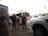 A group of people with backpacks are standing outside, waiting to board or unload a tan minibus. Several parked white vehicles are visible nearby, and some people are holding trolley luggage. The sky is overcast, and there are tall flagpoles in the background.
