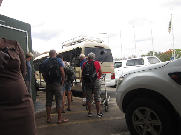 A group of people with backpacks are standing outside, waiting to board or unload a tan minibus. Several parked white vehicles are visible nearby, and some people are holding trolley luggage. The sky is overcast, and there are tall flagpoles in the background.