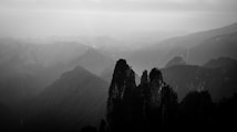 A monochrome image of a mountainous landscape with jagged peaks and layered ridges fading into the mist. The atmosphere is serene and haunting, with the soft interplay of light and shadow creating a dramatic and mysterious effect.