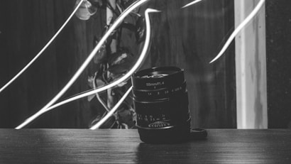 A black and white photo featuring a camera lens sitting on a wooden surface. In the background, there are light streaks and leaves, creating an artistic and abstract visual effect.