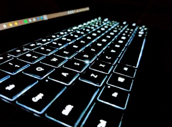 A close-up view of an illuminated keyboard with visible keys arranged in rows. The keys are glowing with a bright white backlight, creating a high contrast against the dark surroundings. The image is angled to capture the depth and alignment of the keyboard, accentuating its sleek and modern design.