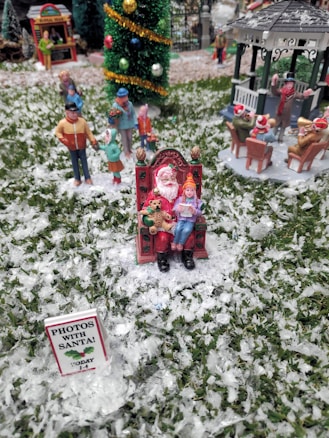 A miniature holiday scene features a figure dressed as Santa Claus sitting on an ornate chair, with a child on their lap, holding a teddy bear. The ground is covered in faux snow, and a sign reads 'Photos with Santa!' in the foreground. In the background, small figurines of people appear to be enjoying a winter setting, including a band playing near a gazebo and a decorated Christmas tree.