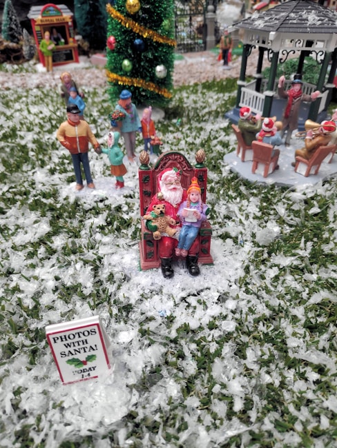 A miniature holiday scene features a figure dressed as Santa Claus sitting on an ornate chair, with a child on their lap, holding a teddy bear. The ground is covered in faux snow, and a sign reads 'Photos with Santa!' in the foreground. In the background, small figurines of people appear to be enjoying a winter setting, including a band playing near a gazebo and a decorated Christmas tree.