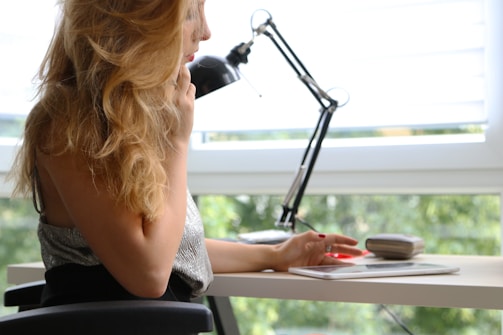 A person with long blond hair is sitting at a desk, holding a phone to their ear. A tablet and a small case or pouch are on the desk. In the background, there is a window with a view of greenery and a black adjustable desk lamp.
