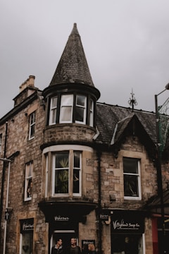A stone building with a prominent conical turret stands under a gray sky. The structure features multiple windows, some with plants visible inside. At the street level, there is a storefront for a shop called 'Highland Soap Co.', and two people are walking in front of it.