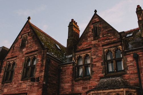 A gothic-style building with red brick walls and ornate arched windows. The roof has steep gables, some parts covered with moss, and chimneys projecting upward. The lighting suggests an evening or early morning setting, providing a warm, slightly eerie atmosphere.