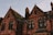 A gothic-style building with red brick walls and ornate arched windows. The roof has steep gables, some parts covered with moss, and chimneys projecting upward. The lighting suggests an evening or early morning setting, providing a warm, slightly eerie atmosphere.