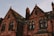 A gothic-style building with red brick walls and ornate arched windows. The roof has steep gables, some parts covered with moss, and chimneys projecting upward. The lighting suggests an evening or early morning setting, providing a warm, slightly eerie atmosphere.