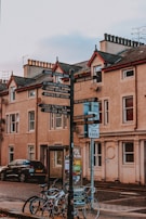 A street view featuring an old-style signpost with multiple directional signs pointing to various local attractions. Surrounding the signpost are bicycles parked against it, a black car, and a background of historic buildings with classic architectural features.
