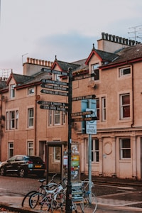 A street view featuring an old-style signpost with multiple directional signs pointing to various local attractions. Surrounding the signpost are bicycles parked against it, a black car, and a background of historic buildings with classic architectural features.