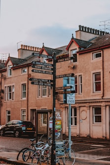 A street view featuring an old-style signpost with multiple directional signs pointing to various local attractions. Surrounding the signpost are bicycles parked against it, a black car, and a background of historic buildings with classic architectural features.