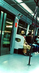 An interior of a subway car with several passengers seated. The environment is modern, featuring metallic and glass elements with overhead lighting. Passengers are dressed in casual clothing; one wears a beige coat and hat. The subway doors are closed, and route maps are visible above.
