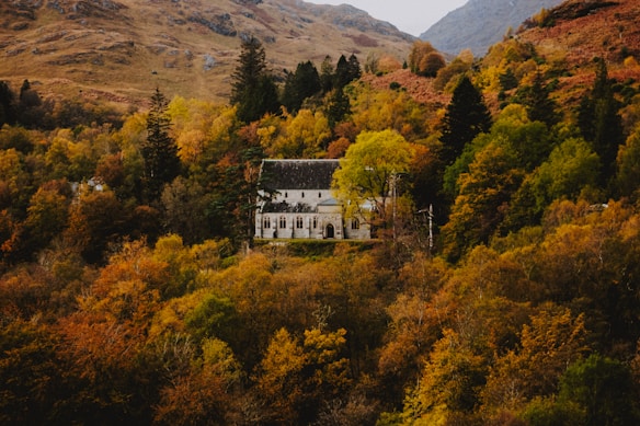 A picturesque white church with a dark roof nestled among a lush landscape of autumn trees. The foliage displays vibrant shades of orange, yellow, green, and brown. It is situated in a mountainous region with gentle hills in the background.