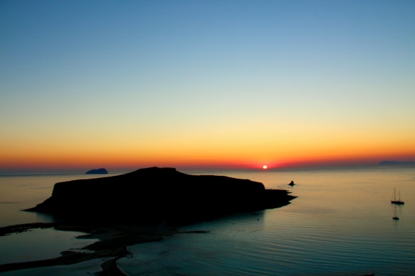A serene seascape at sunset with a vibrant horizon transitioning from deep blue to warm orange and yellow hues as the sun dips below the horizon. Silhouetted landforms, possibly islands, are visible against the colorful sky, and two sailboats are gently anchored in the calm water.