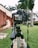A camera mounted on a tripod is positioned on a grassy area. The camera's screen is visible, capturing a building with red and beige walls and surrounded by tall palm trees. The angle suggests a focus on outdoor photography.